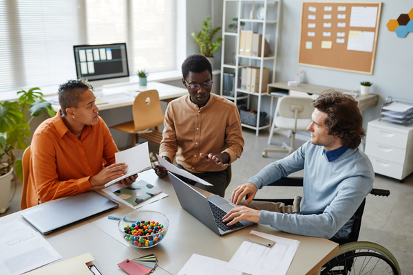 Three people collaborate around a desk in an office, reviewing papers and a laptop