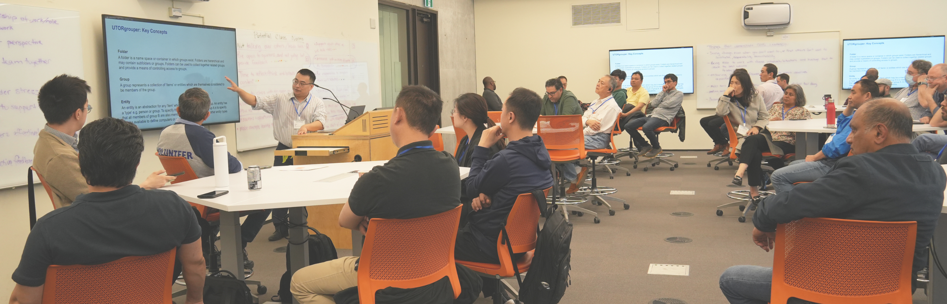 Presenter points to a slide while groups of people sit at tables listen attentively