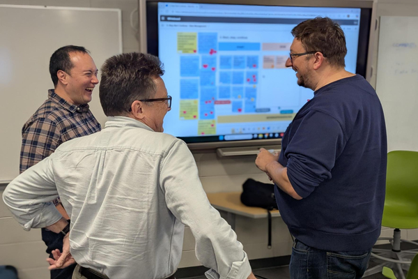 Three people stand and discuss content on a large screen displaying a digital board with notes in a meeting room.